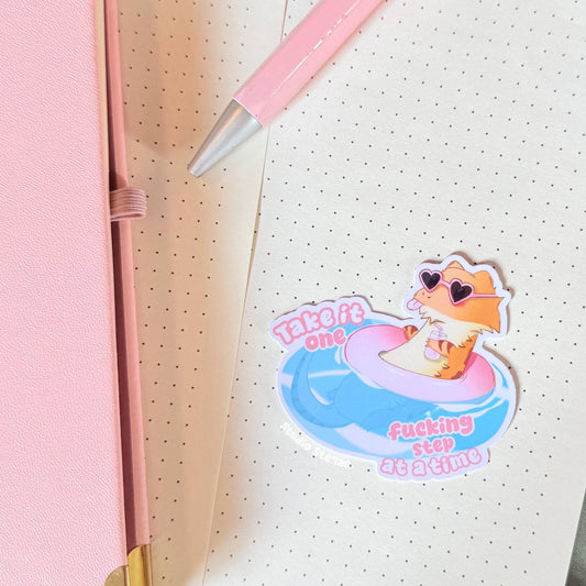 The bearded dragon sticker displayed on a notebook or wall surface in a cozy pastel workspace, showing how the design looks when used for everyday decoration. The floating pink lizard and funny quote add personality and humor to planners and journals. This mental health reminder sticker works great for stationery lovers, students and anyone who enjoys cute reptile art.