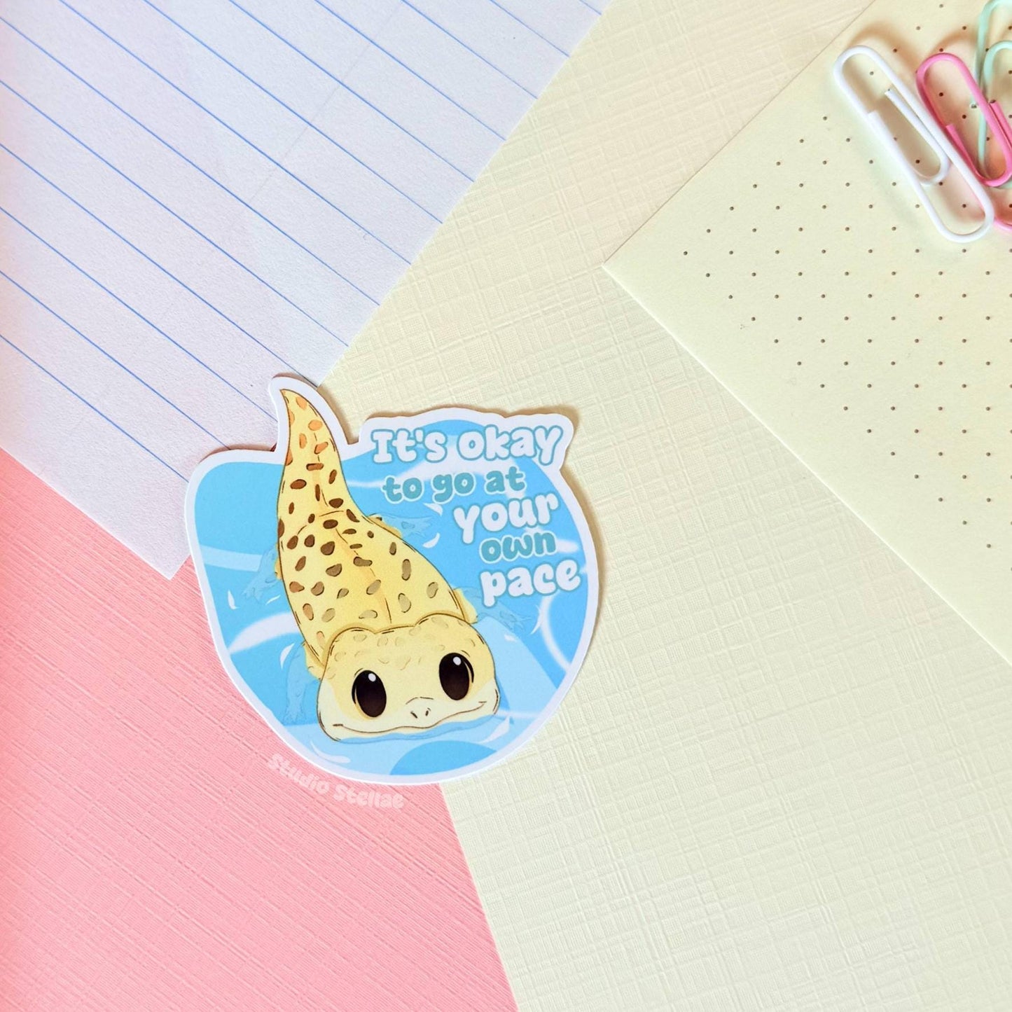 The gecko affirmation sticker displayed on a planner or notebook page in a cozy pastel workspace, showing how the design looks in everyday journaling and stationery setups. The soft colors and gentle quote create a comforting mental health and self care vibe. This motivational reptile decal works perfectly for planners, study notes and laptop decoration.