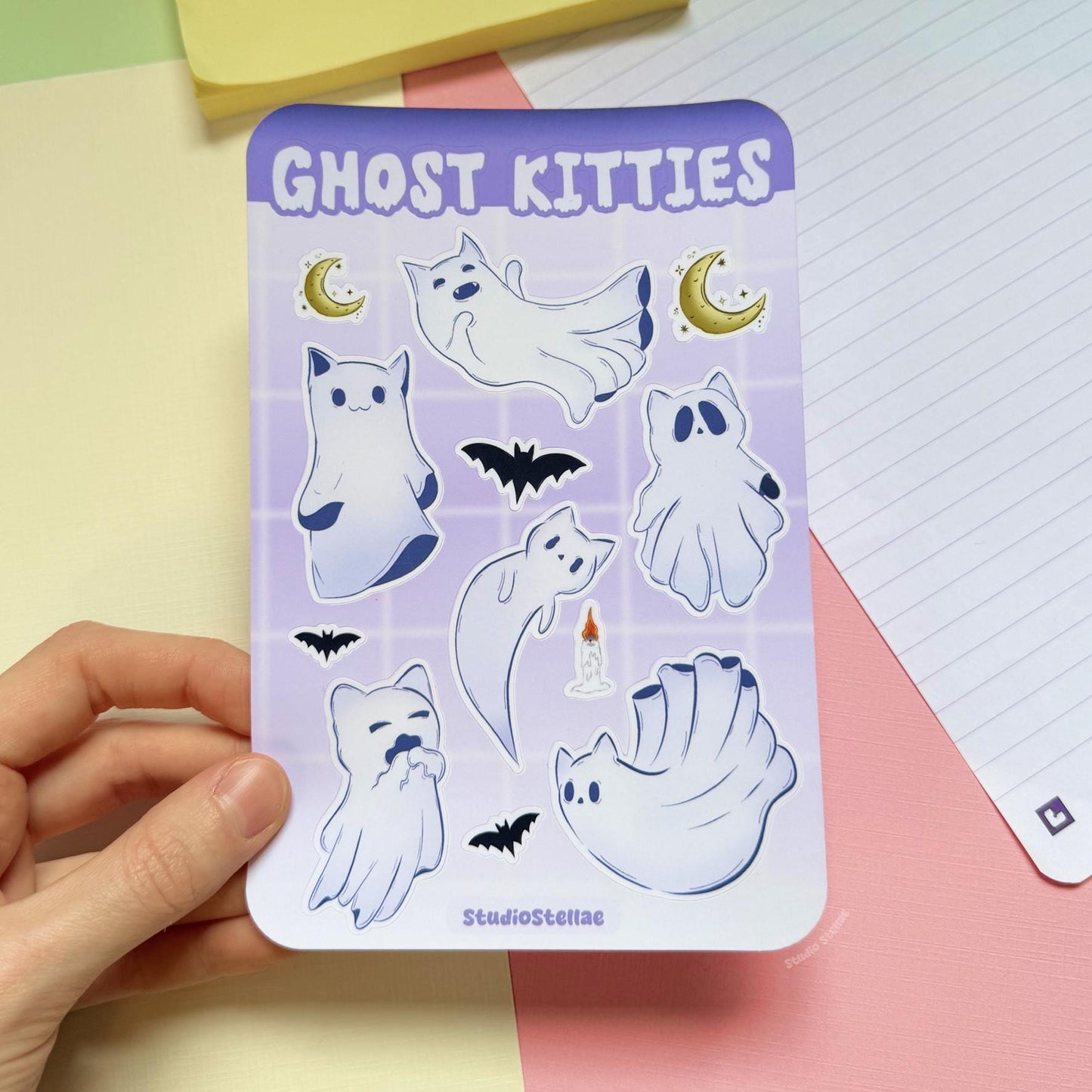 Hand holding the Ghost Kitties sticker sheet above a pastel desk with a notebook in the background. The full sheet shows white ghost cats with soft purple outlines, crescent moons, and small bats in a pastel goth Halloween style. This lifestyle image helps show real-life scale and size in hand, perfect for journaling, planner decorating, scrapbooking, laptops, and adding spooky witchy vibes to everyday surfaces.