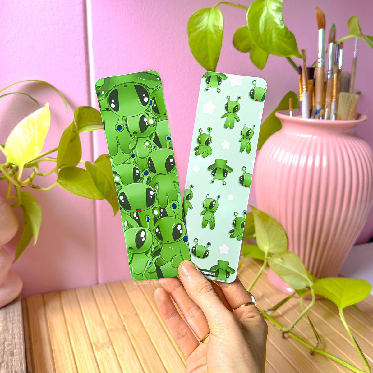 Hand holding the green alien bookmark set in a cozy pastel workspace with a pink wall, leafy green plants, and a pink vase filled with paintbrushes. Both bookmarks are visible, showing their bright green alien designs and smooth matte lamination. This lifestyle photo shows real-life scale in hand and how the bookmarks look in natural light, perfect for book lovers, journaling, school supplies, and gifting.
