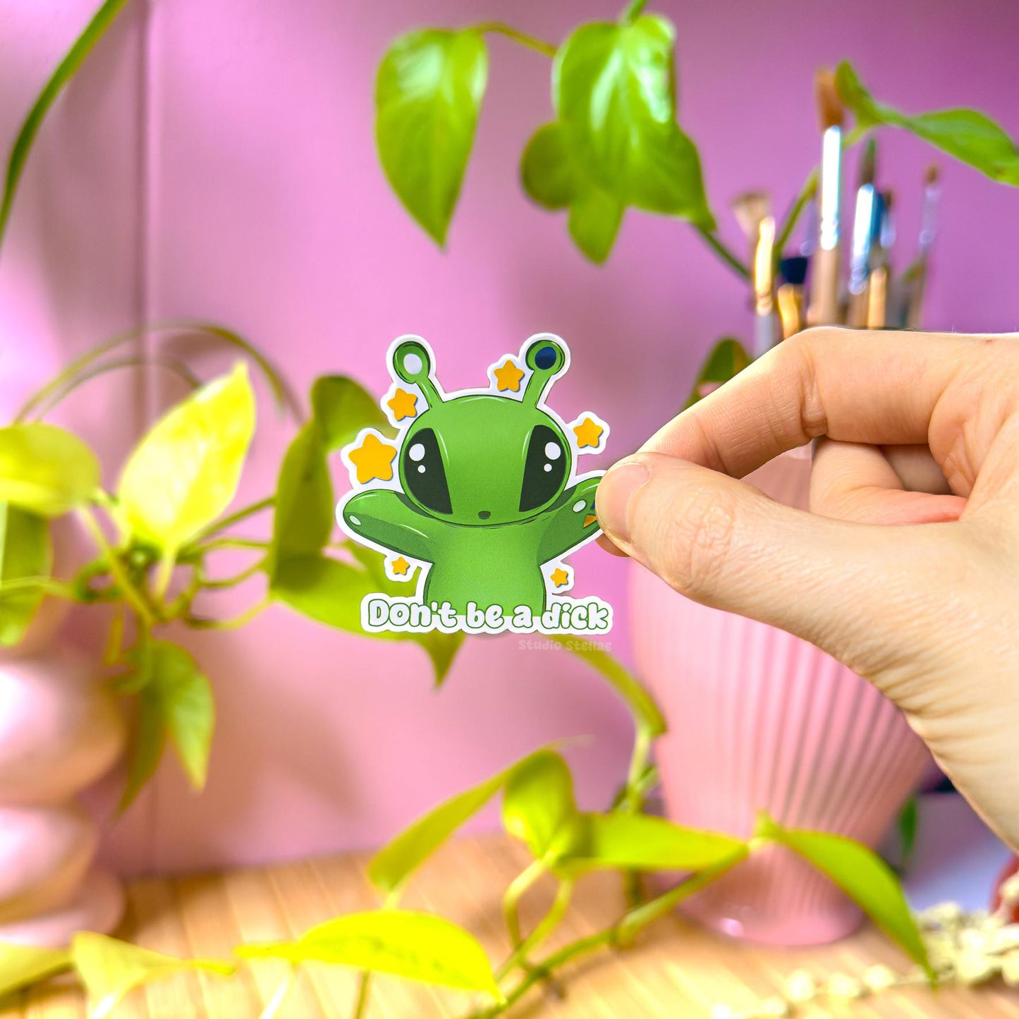 Close-up of a hand peeling and applying the Aftonsparv Alien Sticker beside a small plant to show scale, flexibility and thickness. The matte laminated surface and clean die cut edges highlight the durable, water resistant finish. This handmade IKEA plushie fan art decal and Don’t Be a Dick funny quote sticker is easy to place on laptops, journals, water bottles and planners for everyday cute alien decoration.
