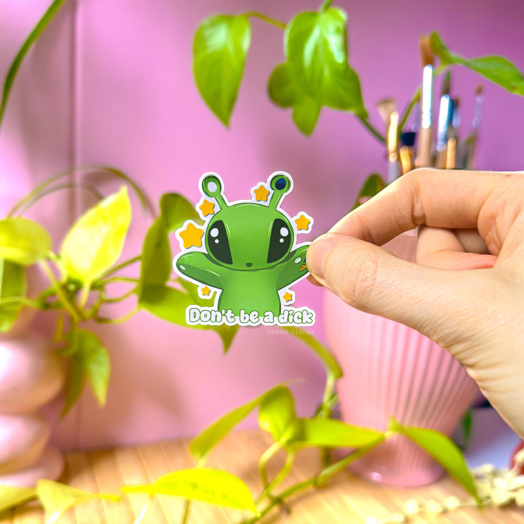 Close-up of a hand peeling and applying the Aftonsparv Alien Sticker beside a small plant to show scale, flexibility and thickness. The matte laminated surface and clean die cut edges highlight the durable, water resistant finish. This handmade IKEA plushie fan art decal and Don’t Be a Dick funny quote sticker is easy to place on laptops, journals, water bottles and planners for everyday cute alien decoration.