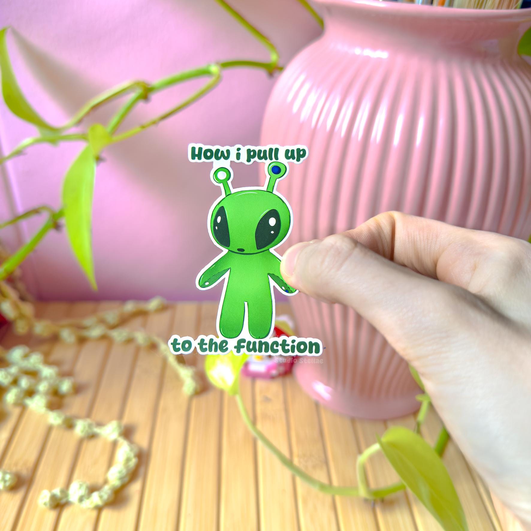 Close-up lifestyle photo of a hand peeling and placing the Aftonsparv alien sticker near a small plant to show size and flexibility. The matte laminated surface and clean die cut edges highlight the durable, water resistant finish. This cute chibi IKEA alien fan art decal works easily on laptops, water bottles, notebooks and journals, making it a practical and fun funny quote sticker for everyday use.