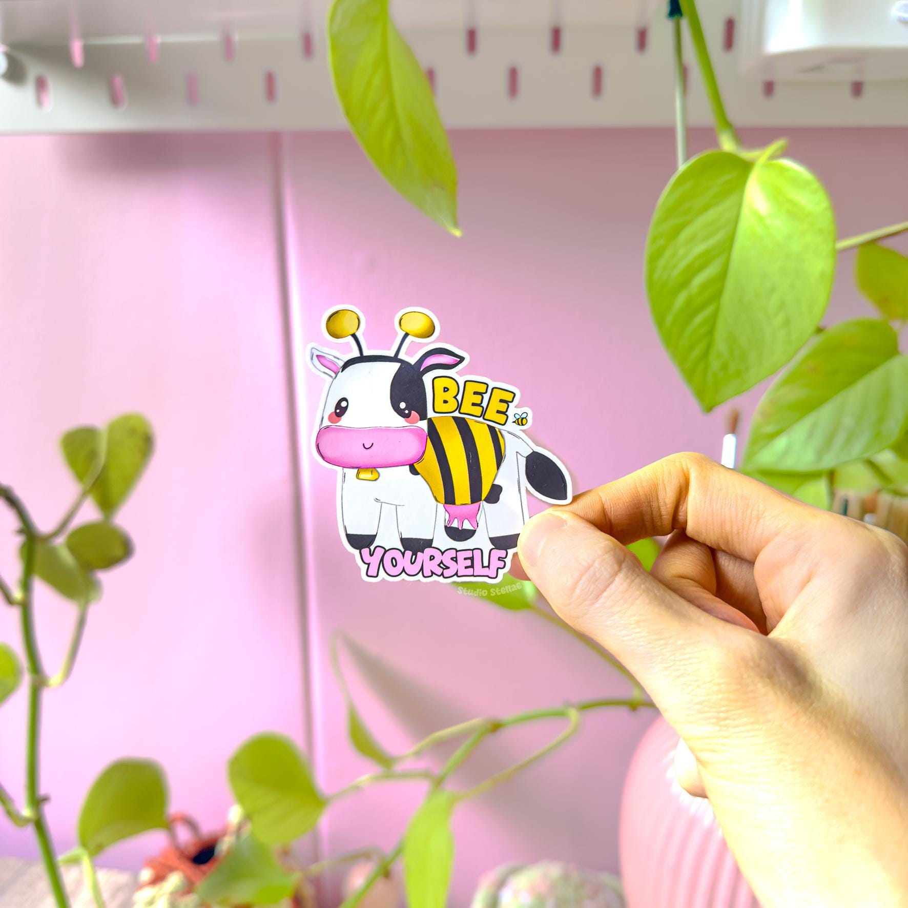 Close-up of a hand peeling and placing the Bee Yourself cow sticker onto a smooth surface near a plant to show size and flexibility. The matte laminated surface and clean die cut edges highlight the durable, water resistant finish. This cute bumblebee cow decal applies easily to laptops, water bottles, planners and journals, making it a fun and uplifting affirmation sticker for everyday positivity.