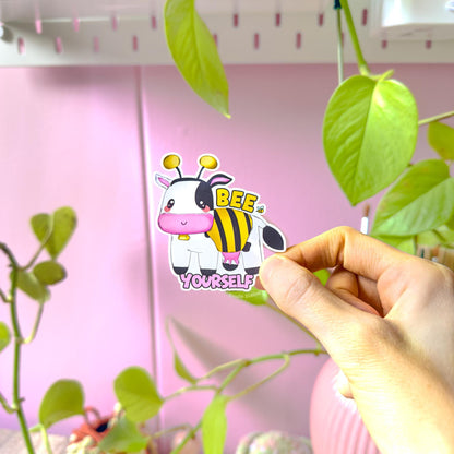 Close-up of a hand peeling and placing the Bee Yourself cow sticker onto a smooth surface near a plant to show size and flexibility. The matte laminated surface and clean die cut edges highlight the durable, water resistant finish. This cute bumblebee cow decal applies easily to laptops, water bottles, planners and journals, making it a fun and uplifting affirmation sticker for everyday positivity.