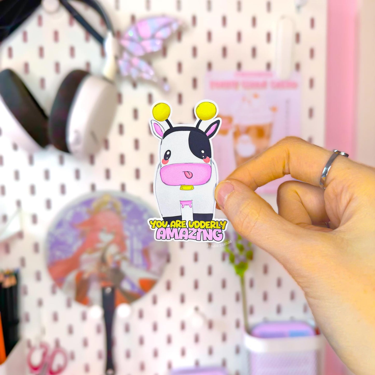 Close-up of a hand peeling and placing the Cow Bumblebee sticker onto a smooth surface near a plant to show size and flexibility. The matte laminated surface and clean die cut edges highlight the durable finish. This cute pink and yellow bee cow decal applies easily to journals, planners, laptops and water bottles, making it a sweet everyday positivity reminder.