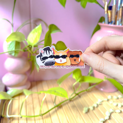 Close-up of a hand peeling and placing the animal party sticker onto a smooth surface near a small plant to show size and flexibility. The matte laminated surface and clean die cut edges highlight the durable finish. This colorful chibi cat, dog and zebra decal applies easily to journals, planners, laptops and water bottles, making it a fun dark humor and mental health coping sticker for everyday use.