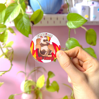 Lifestyle photo of the ferret chaos sticker held up in front of bright green plants, highlighting the glossy colors and bold die cut shape. The kawaii ferret sits confidently on a chair with flames and the text “Embrace the Chaos,” giving cute-but-unhinged meme vibes. Handmade matte laminated decal designed for water bottles, laptops, journals and planners, a fun gift for ferret lovers and anyone who relates to chaotic humor.