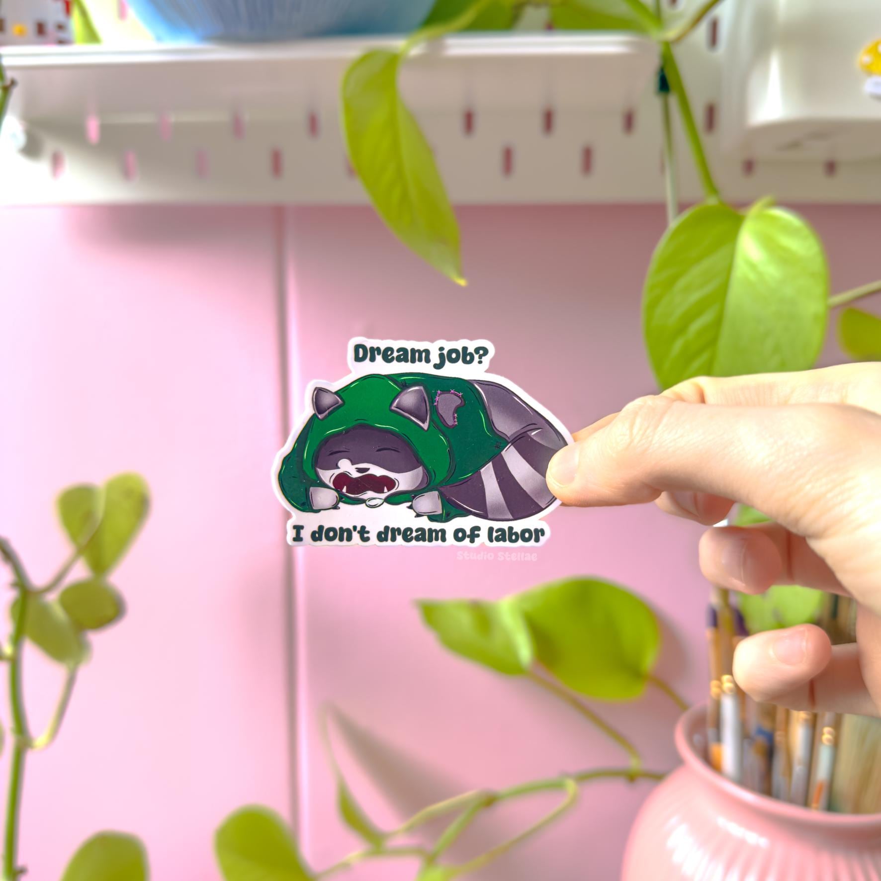 Close-up of a hand holding the raccoon sticker near green plant leaves in natural light, showing true size and color detail. The sleepy kawaii trash panda design and sarcastic anti-work quote create a soft but rebellious vibe. Perfect as a laptop decal, planner sticker or water bottle decoration for fans of relatable humor.