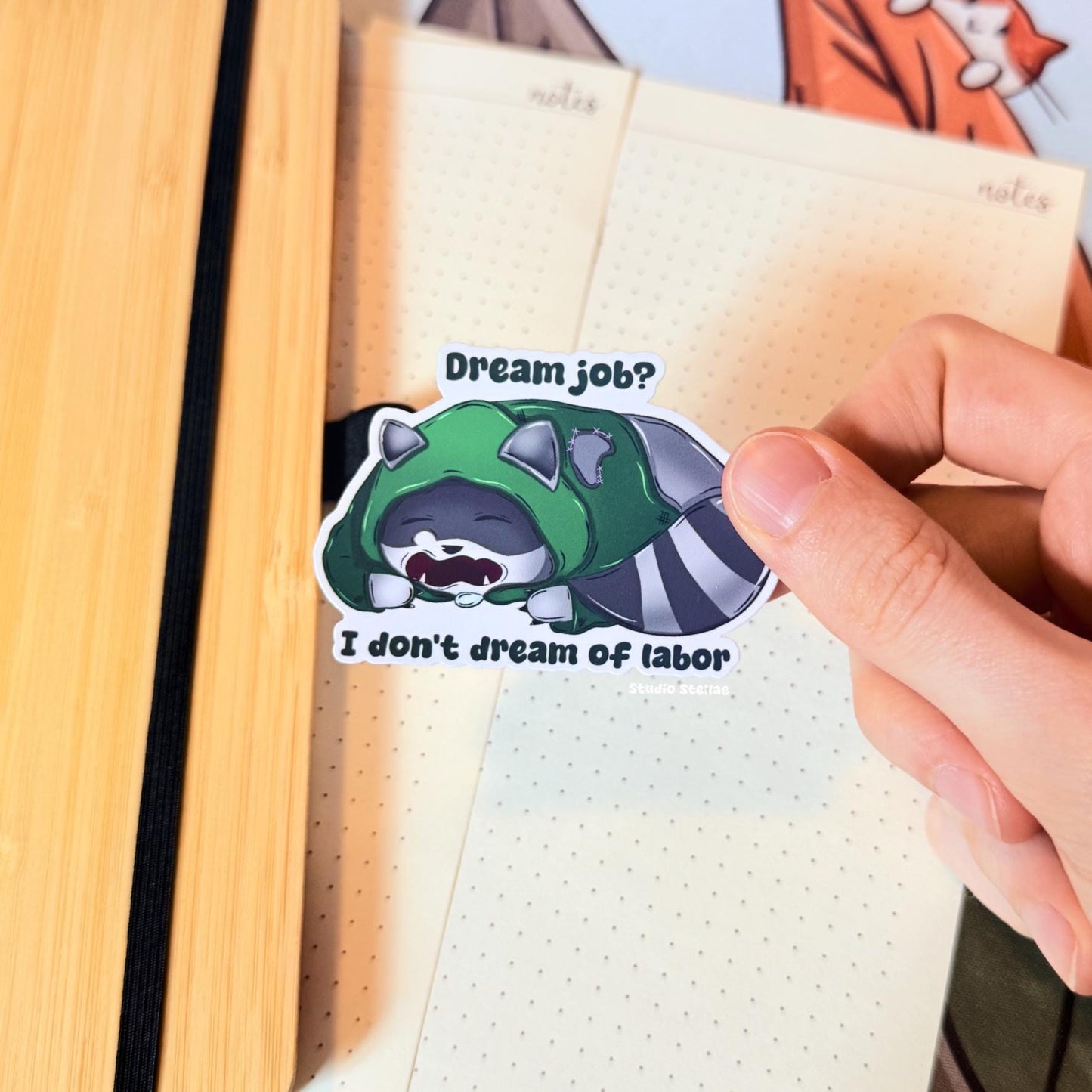 The rogue raccoon sticker placed on a notebook page to show scale and everyday use. The tired trash panda lies down beneath the quote “Dream Job? I Don’t Dream of Labor,” adding relatable burnout and anti-work humor to journals and planners. This matte laminated vinyl decal works great on notebooks, laptops and water bottles.
