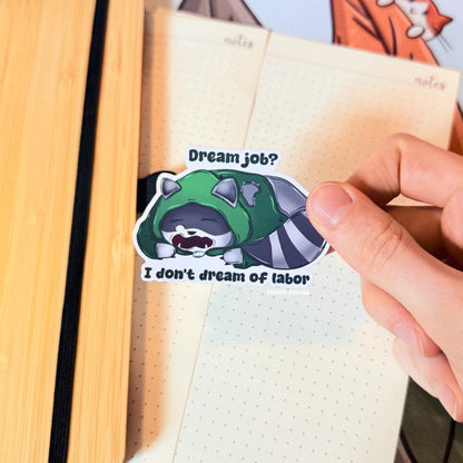 The rogue raccoon sticker placed on a notebook page to show scale and everyday use. The tired trash panda lies down beneath the quote “Dream Job? I Don’t Dream of Labor,” adding relatable burnout and anti-work humor to journals and planners. This matte laminated vinyl decal works great on notebooks, laptops and water bottles.