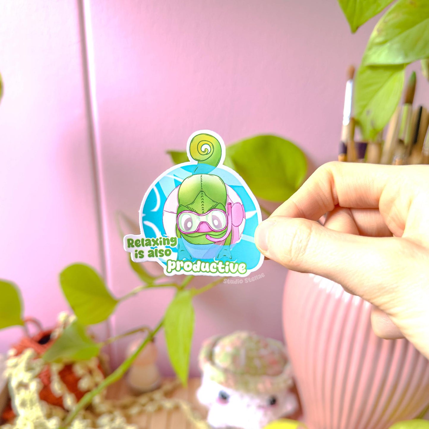 Close-up of a hand peeling and placing the chameleon sticker onto a smooth surface near a plant to show size and flexibility. The matte laminated surface and clean die cut edges highlight the durable finish. This cute green lizard decal applies easily to journals, planners, laptops and water bottles, making it a calming everyday self care and relax reminder.