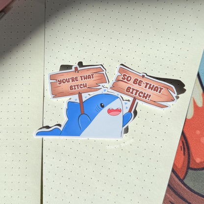 The cute shark sticker displayed on a notebook or planner page in a cozy pastel workspace, showing how the design looks in everyday stationery setups. The bold “You’re that bitch” quote adds confident motivational humor to study notes and journals. This self love shark decal works perfectly for planners, laptops and water bottles.