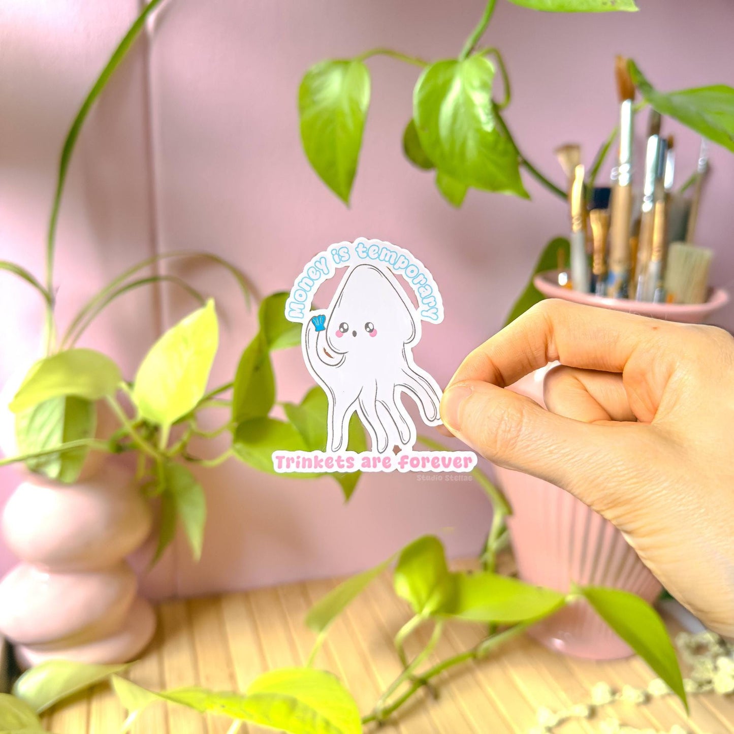 Close-up of a hand holding the white squid sticker near green plants in natural light to show true size and color. The soft pastel ocean creature and “Money is temporary, trinkets are forever” quote create playful anti-capitalist humor in a kawaii style. This matte laminated decal is great for laptops, planners, notebooks and water bottles as a cute sea sticker and trinket lover reminder.