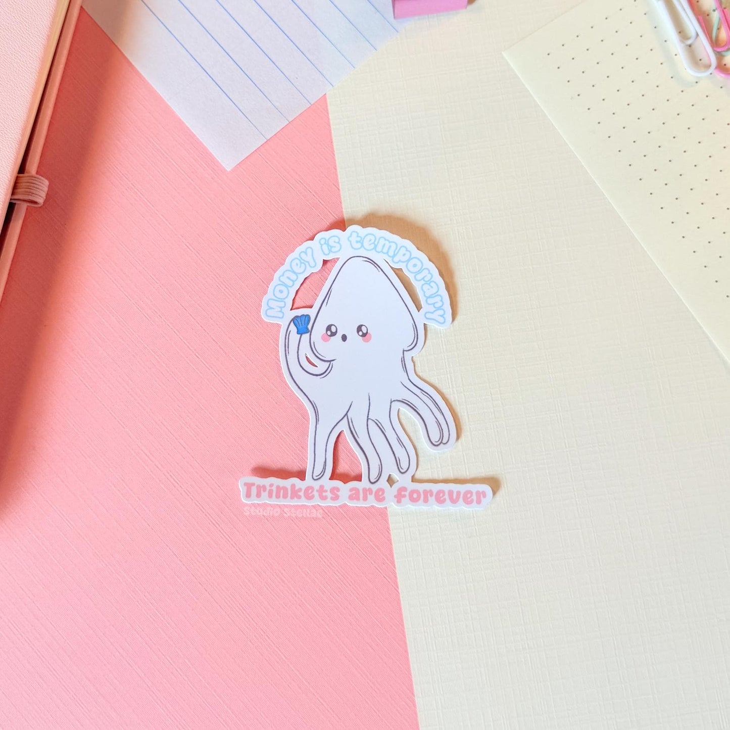 White squid sticker displayed on a notebook or paper surface with pastel stationery layers, showing how it looks when used for journaling and planning. The cute squid and “Money is temporary, trinkets are forever” quote add playful humor to planners and notebooks. This handmade matte laminated vinyl sticker has clean die cut edges and works perfectly for journals, laptops and water bottles.