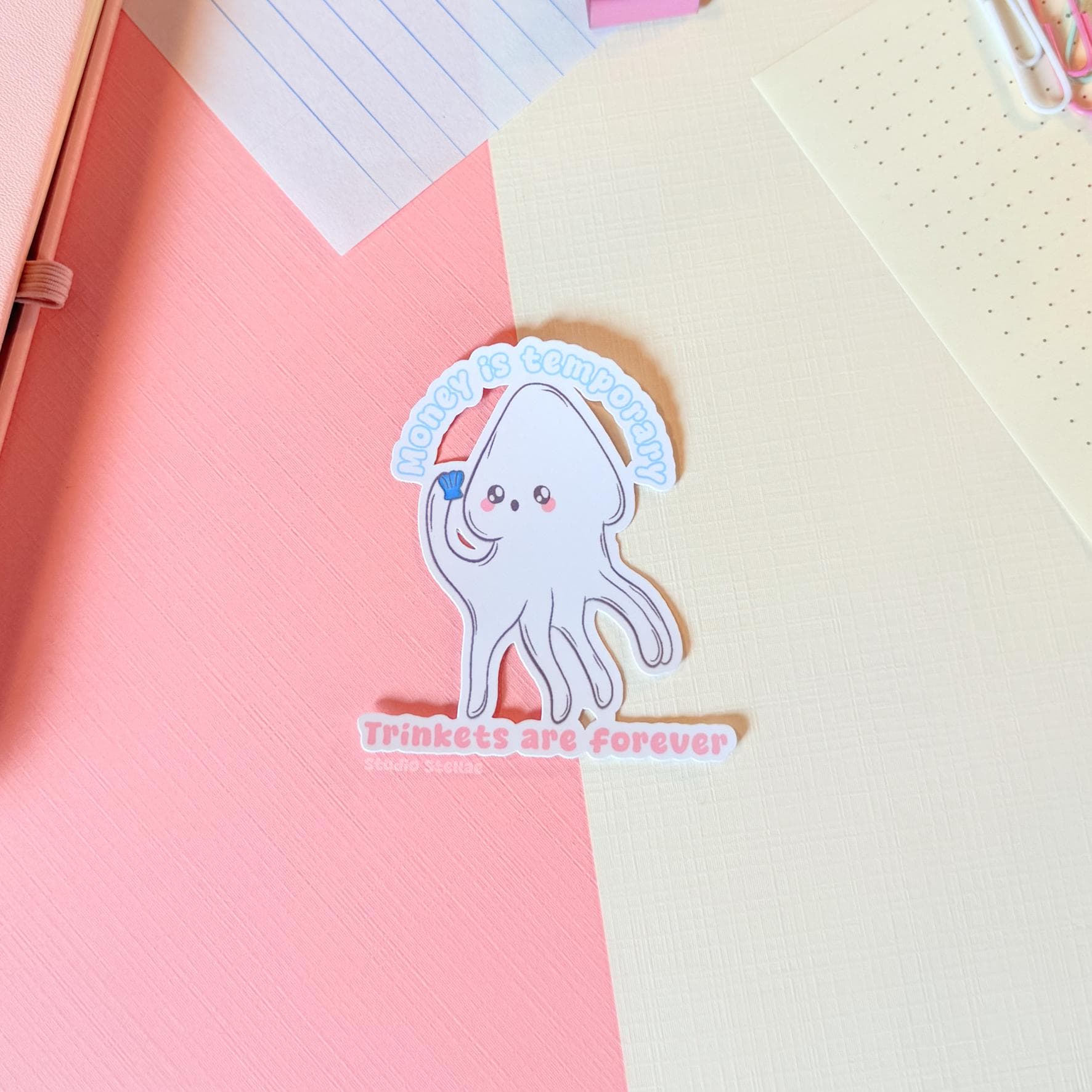 White squid sticker displayed on a notebook or paper surface with pastel stationery layers, showing how it looks when used for journaling and planning. The cute squid and “Money is temporary, trinkets are forever” quote add playful humor to planners and notebooks. This handmade matte laminated vinyl sticker has clean die cut edges and works perfectly for journals, laptops and water bottles.