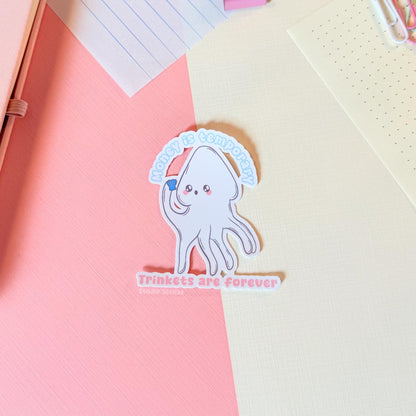 White squid sticker displayed on a notebook or paper surface with pastel stationery layers, showing how it looks when used for journaling and planning. The cute squid and “Money is temporary, trinkets are forever” quote add playful humor to planners and notebooks. This handmade matte laminated vinyl sticker has clean die cut edges and works perfectly for journals, laptops and water bottles.