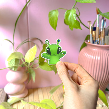 Close-up of a hand holding the Aftonsparv alien sticker with raised arms near houseplants, showing real-life scale, smooth matte finish and crisp die cut outline. The plushie inspired green alien design looks bright and cheerful against a pastel background. Ideal as a cute laptop decal, planner sticker or journal decoration for fans of kawaii IKEA alien fan art and handmade stationery.