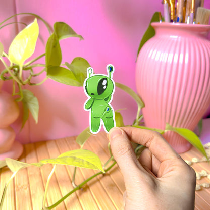 Lifestyle shot of a hand placing an Aftonsparv alien sticker on a surface near a pink vase and leafy plants, creating a cozy desk scene that shows how the decal looks in real use. The cheerful green kawaii alien adds playful charm to planners, scrapbooks, laptops or water bottles. Demonstrates size, color accuracy and the soft matte laminated finish in natural lighting.