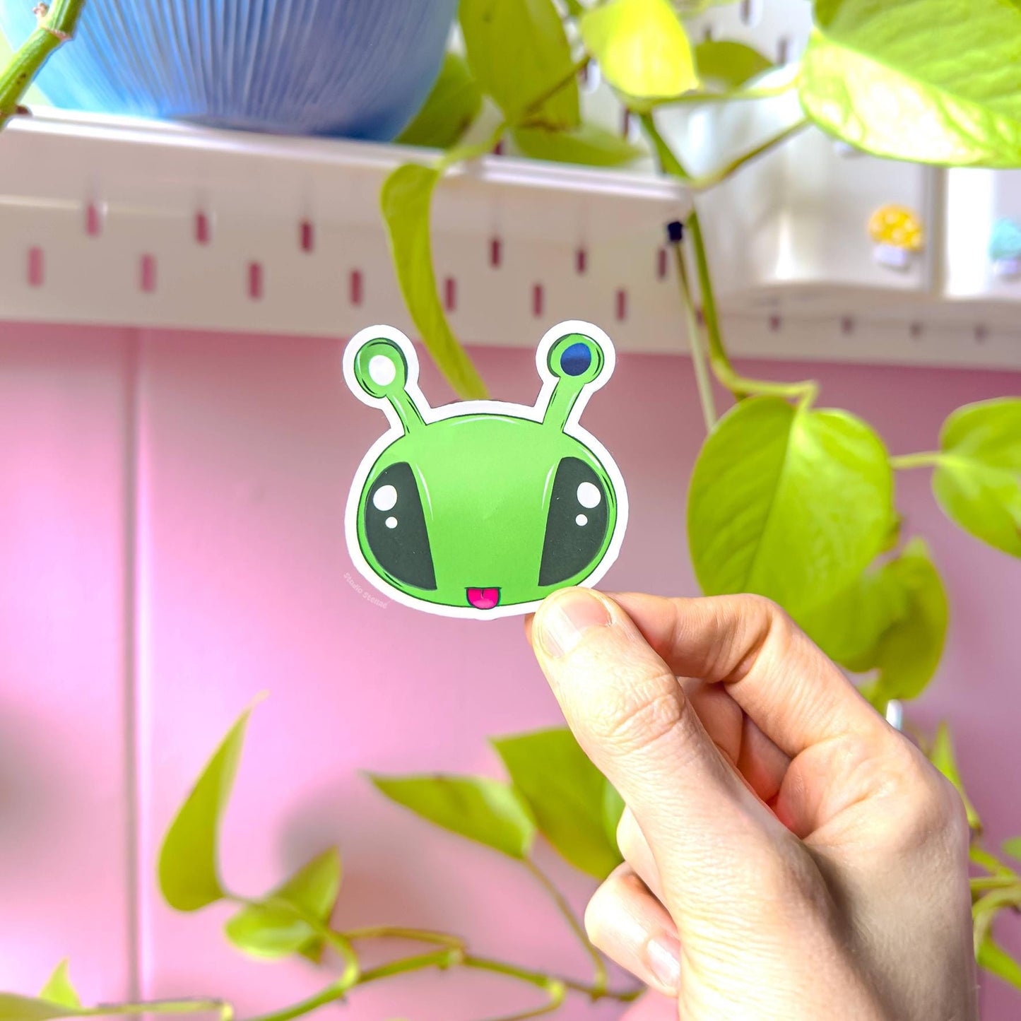 Hand holding the wide alien face sticker with large eyes and antennas against a pastel pink background, clearly showing bright colors and smooth matte texture. This plushie inspired Aftonsparv design has a bold, expressive look that stands out on laptops, notebooks and planners. A cute kawaii alien decal that adds fun personality to everyday stationery and accessories.