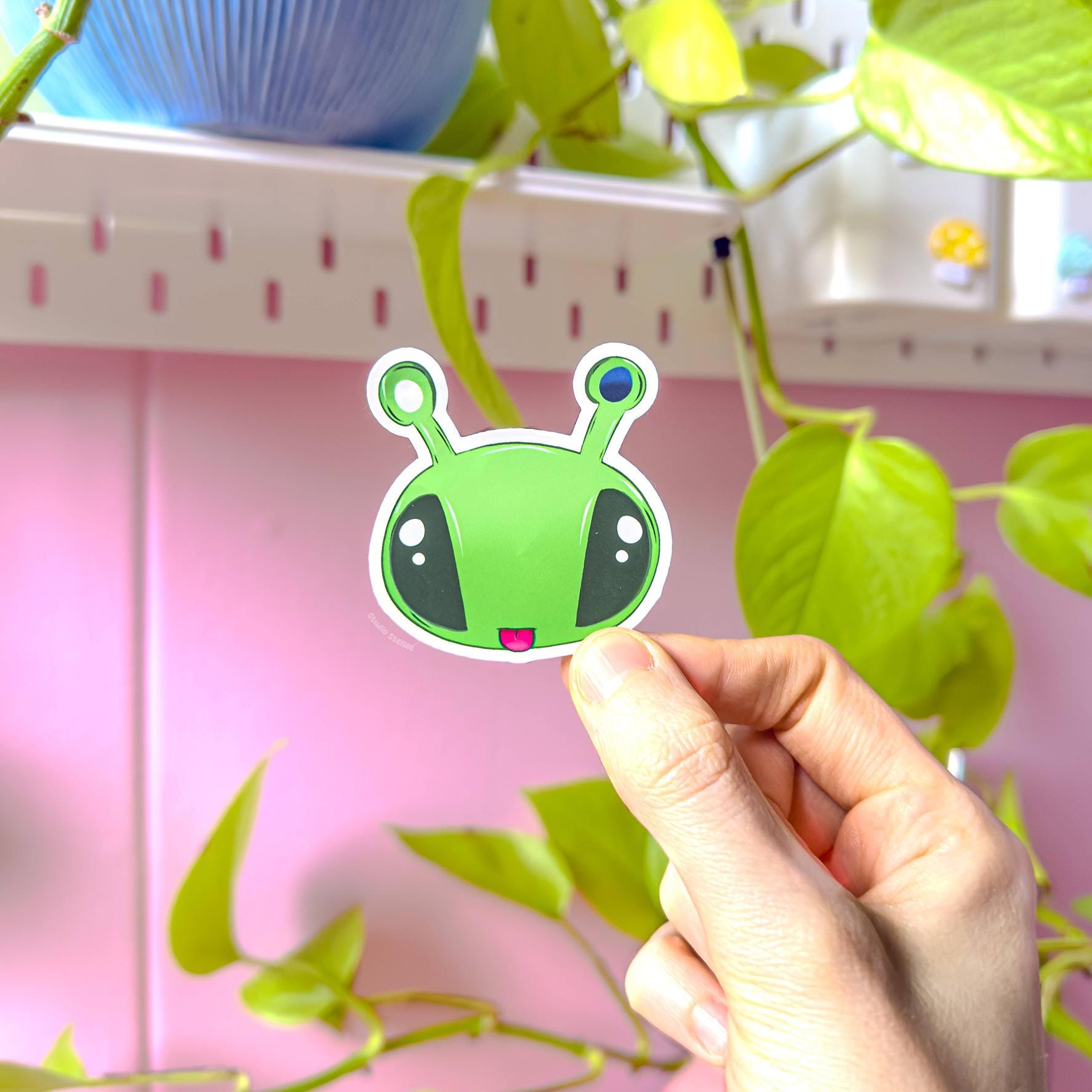 Hand holding the wide alien face sticker with large eyes and antennas against a pastel pink background, clearly showing bright colors and smooth matte texture. This plushie inspired Aftonsparv design has a bold, expressive look that stands out on laptops, notebooks and planners. A cute kawaii alien decal that adds fun personality to everyday stationery and accessories.