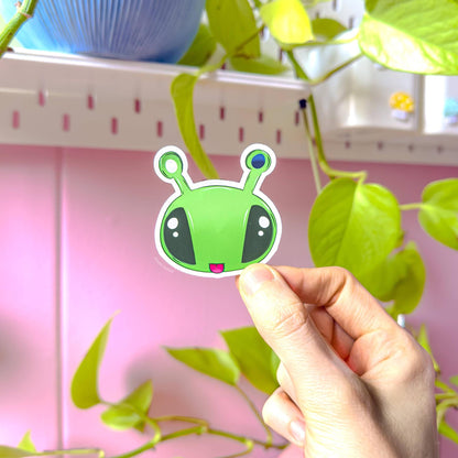 Hand holding the wide alien face sticker with large eyes and antennas against a pastel pink background, clearly showing bright colors and smooth matte texture. This plushie inspired Aftonsparv design has a bold, expressive look that stands out on laptops, notebooks and planners. A cute kawaii alien decal that adds fun personality to everyday stationery and accessories.