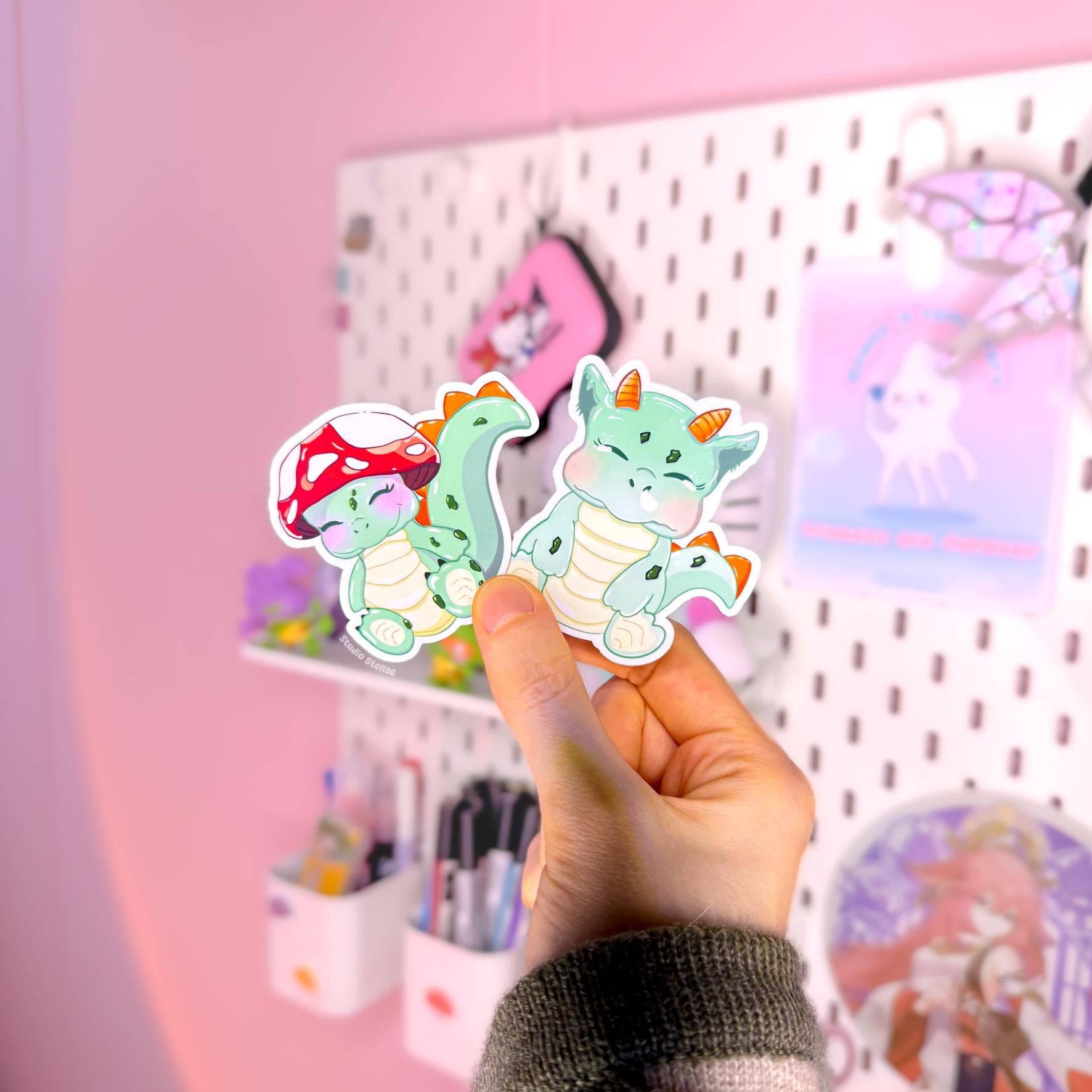 Hand holding both baby dinosaur stickers together in soft natural light, showing their true size, matte texture and clean die cut edges. The sleepy mint green dino and the tiny mushroom hat friend create a cute besties vibe that feels cozy and playful. Perfect for decorating planners, journals, laptops and water bottles with sweet cottagecore fantasy creature charm.