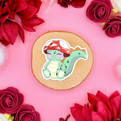 Close-up of the sleepy baby dinosaur sticker placed on cork with red flowers, highlighting soft pastel colors, rounded chibi features and smooth matte laminate. The calm expression and gentle pose give this kawaii dino a cozy cottagecore feel. A durable waterproof decal perfect for planners, scrapbooks, laptops and water bottles for fans of cute stationery and fantasy creatures.
