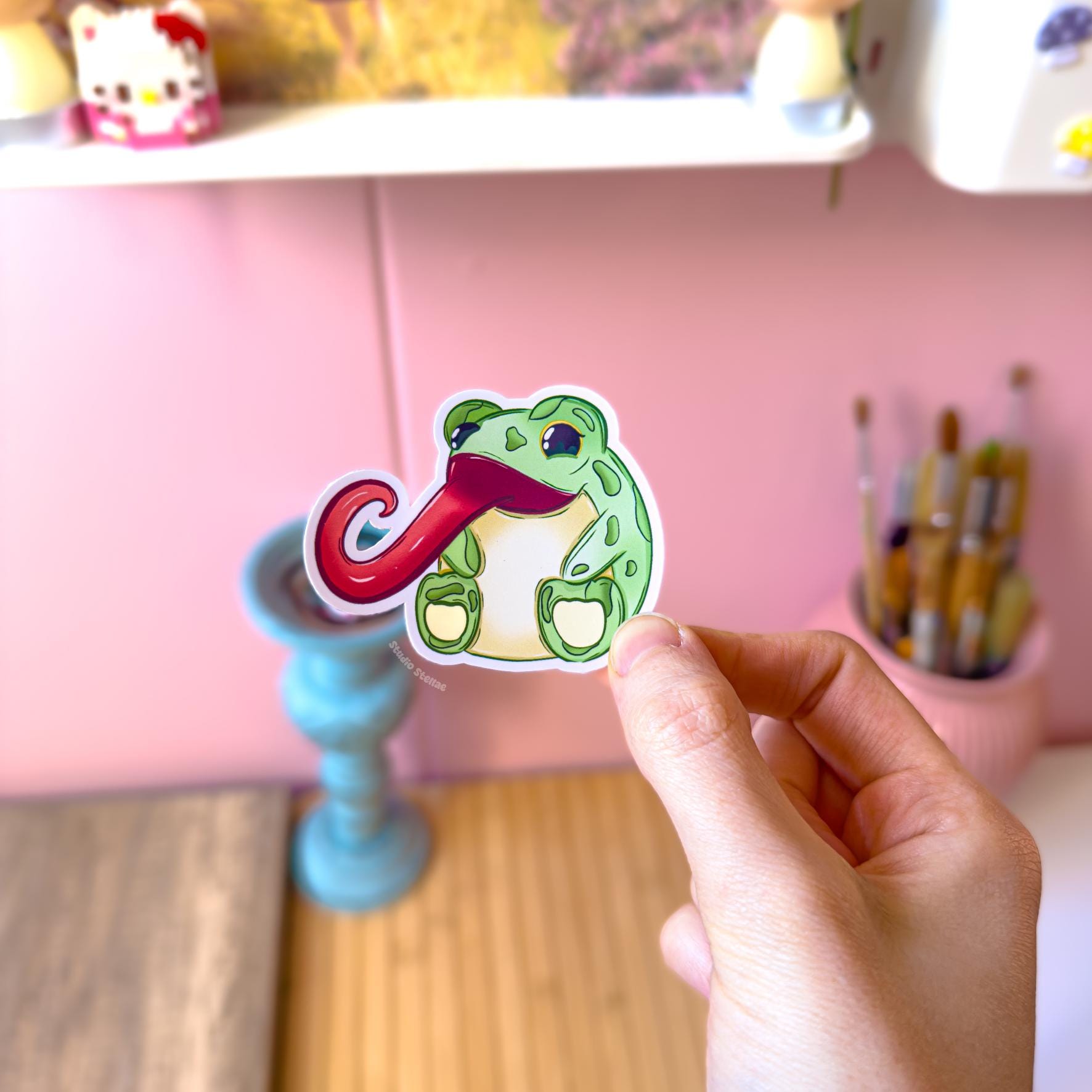 Close-up of the frog with a ridiculously long pink tongue held between fingers, photographed against a pastel desk background to show detail and scale. The exaggerated expression and goofy pose give this sticker lots of personality. Waterproof and matte laminated, it’s perfect as a funny laptop decal, planner sticker or water bottle decoration.