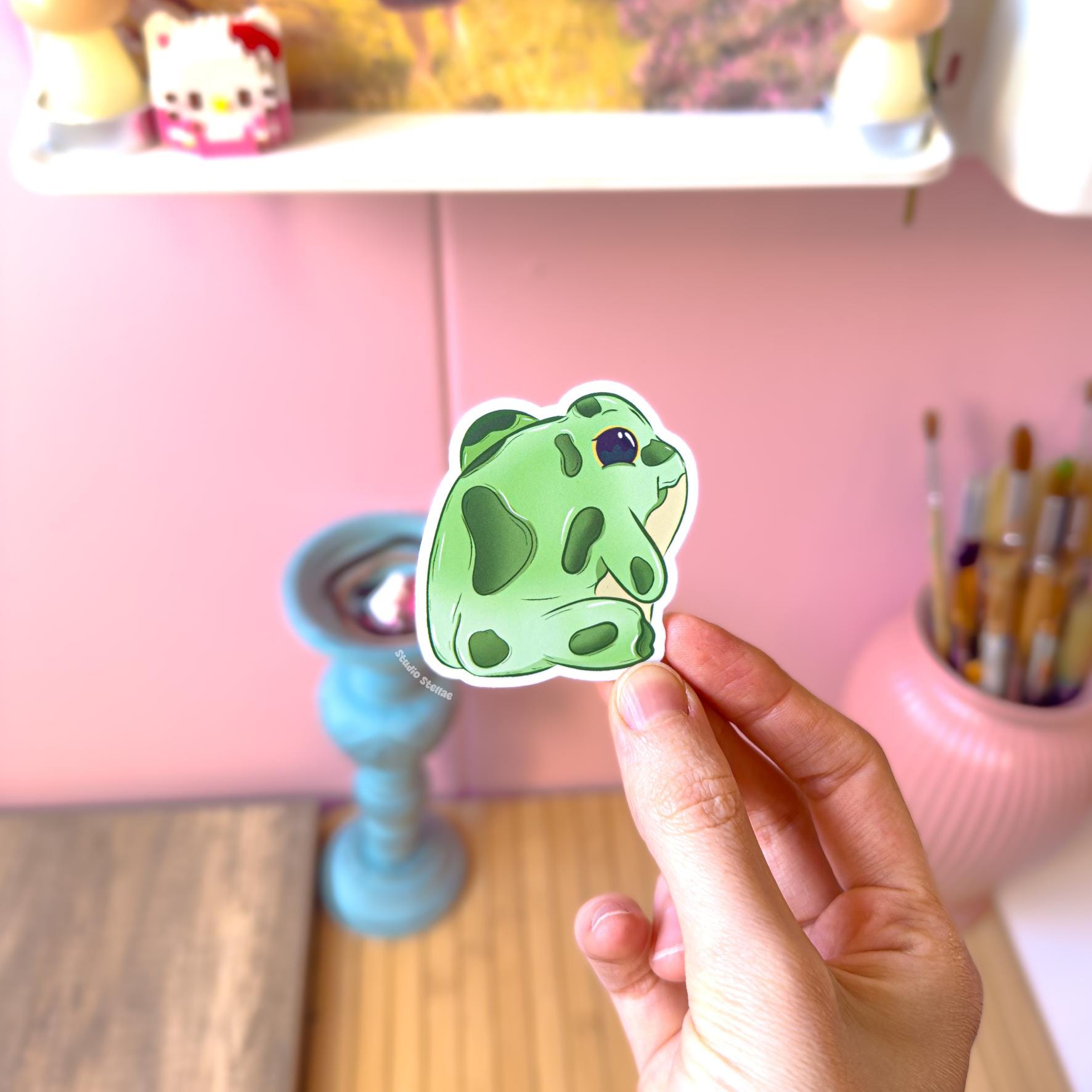 Close-up of a hand holding the frog bum sticker, highlighting the rounded green shape, tiny legs and cute smiling face on the back. The smooth matte laminate protects against scratches and water while keeping colors vibrant. A silly kawaii frog decal that adds humor to planners, scrapbooks, laptops and water bottles.