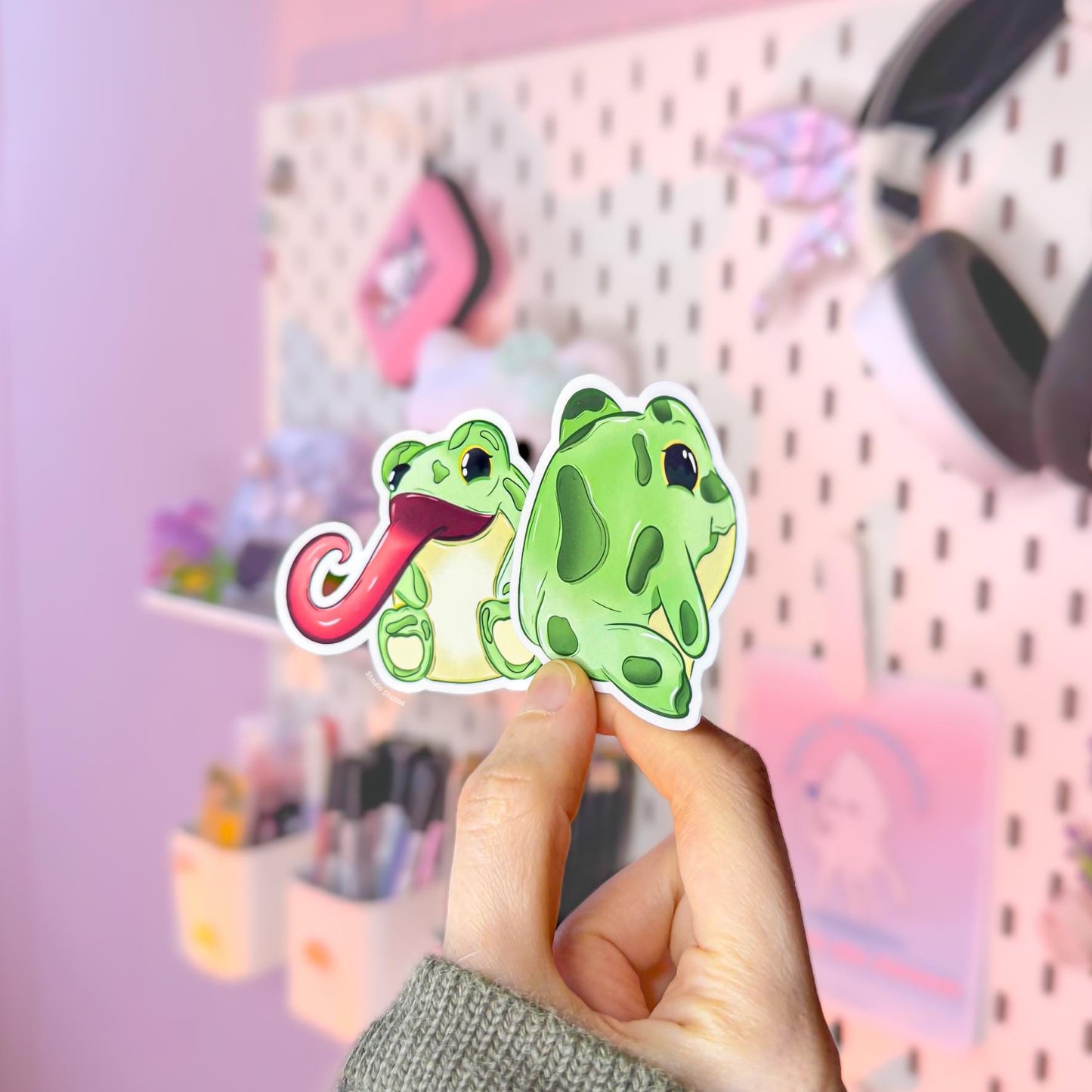Hand holding both frog stickers together in soft natural light, showing their real-life size, matte texture and crisp die cut edges. The long tongue frog and cheeky frog bum design create a funny best-friends duo with lots of personality. Perfect for decorating planners, journals, laptops and water bottles with playful green animal charm.