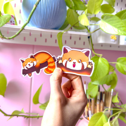 Two hands holding both red panda stickers side by side against a pastel studio background with plants and desk decor. The photo shows true size and scale, letting customers see how the happy and sleepy pandas look when applied to real objects. The matte finish and clean die-cut edges are visible, helping buyers imagine decorating planners, laptops, water bottles and notebooks with these cute animal decals.
