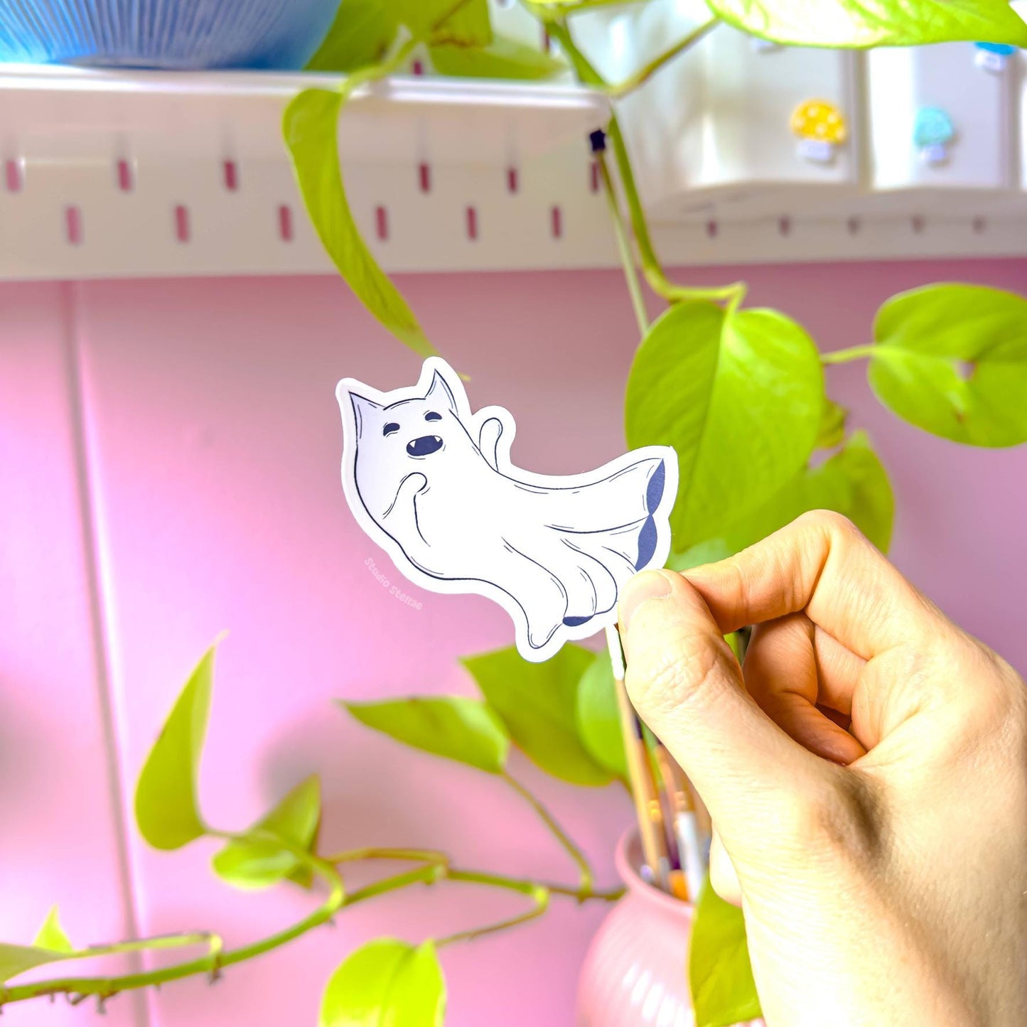 Close-up of a single ghost kitty sticker gently held between fingers near leafy houseplants and a soft pink studio wall. The floating pastel cat design looks playful and slightly spooky, blending kawaii cuteness with a subtle pastel goth mood. This shot highlights the smooth matte laminate, bright print clarity, and precise die-cut outline. A charming Halloween decal perfect for adding personality to planners, journals, water bottles, and cozy stationery setups.