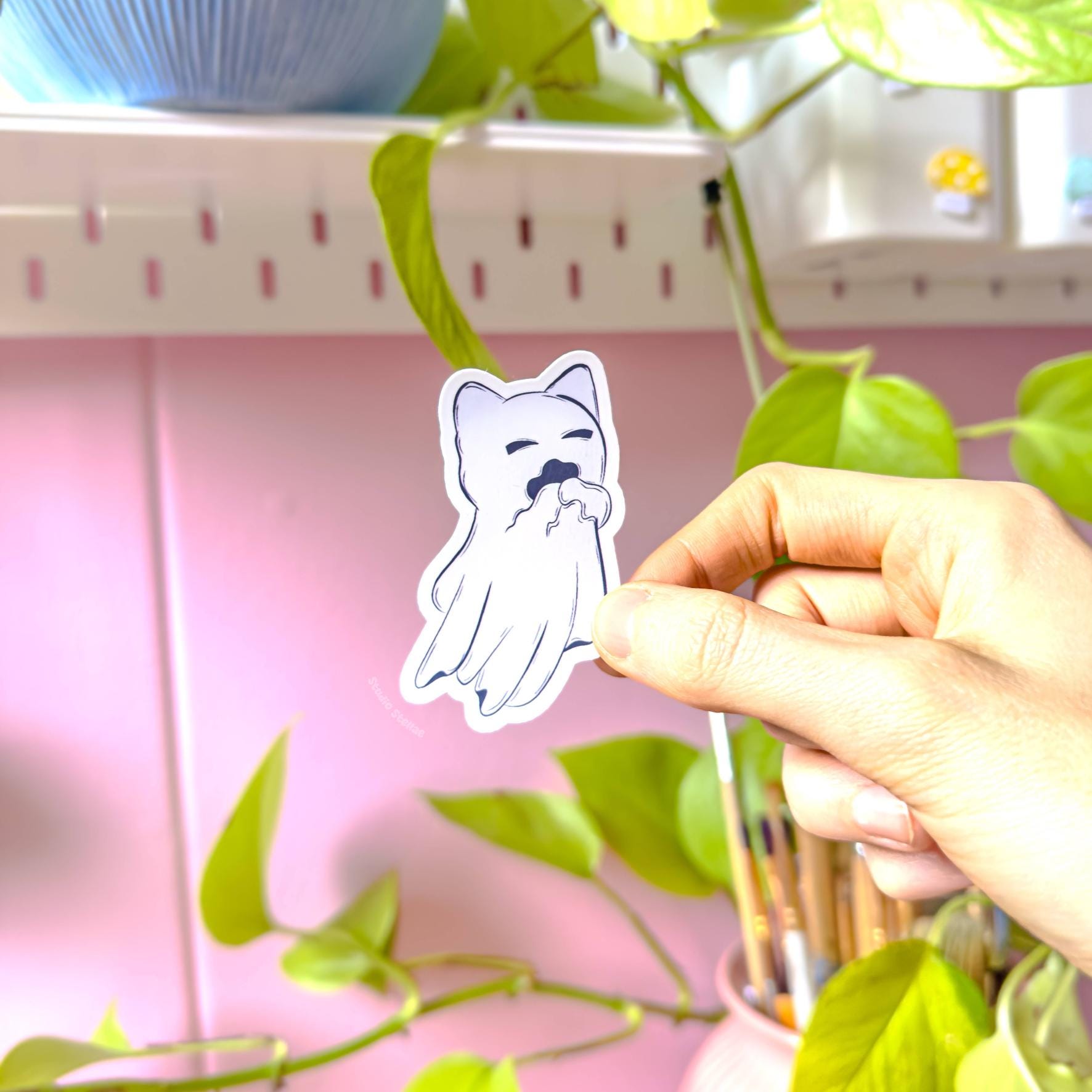 Close-up of a single pastel goth ghost cat sticker being held between fingers near hanging plants, photographed in warm daylight. The floating kitty has a different pose and expression than the other designs, showing the variety in the Ghost Kitty sticker set. The image highlights clean linework, sturdy sticker paper, and a silky matte laminated finish that protects the print. Cute spooky Halloween cat art decal for decorating journals, scrapbooks, phones, and cozy stationery.