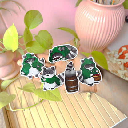 Rogue raccoon sticker set displayed near a pink desk and houseplant, showing several trash panda rogue characters grouped together. This lifestyle photo highlights real life scale, matte finish, and soft colors of the fantasy raccoon decals, demonstrating how they look when used to decorate planners, journals, laptops, or tabletop gaming accessories while adding cute DnD inspired personality to everyday spaces.