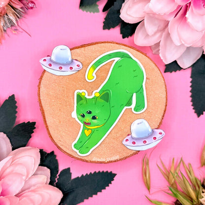 Close up of the long stretching alien cat sticker on a pink background with flowers and small UFO props, showing the full shape of the green cosmic kitty design. This image emphasizes vibrant color, crisp outline, and matte waterproof finish of the kawaii space cat decal. Perfect for cat sticker collectors, sci fi lovers, and anyone decorating a laptop, water bottle, planner, or journal with slightly weird but adorable galaxy themed stationery.