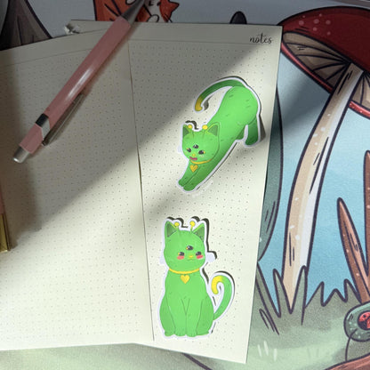 Alien cat sticker applied to a notebook page near drawing tools, showing how the green cosmic kitty decal looks when used in real life. Highlights scale, matte finish, and adhesion, demonstrating that this kawaii space cat sticker works beautifully for planners, journals, sketchbooks, or creative spaces while adding playful sci fi personality.