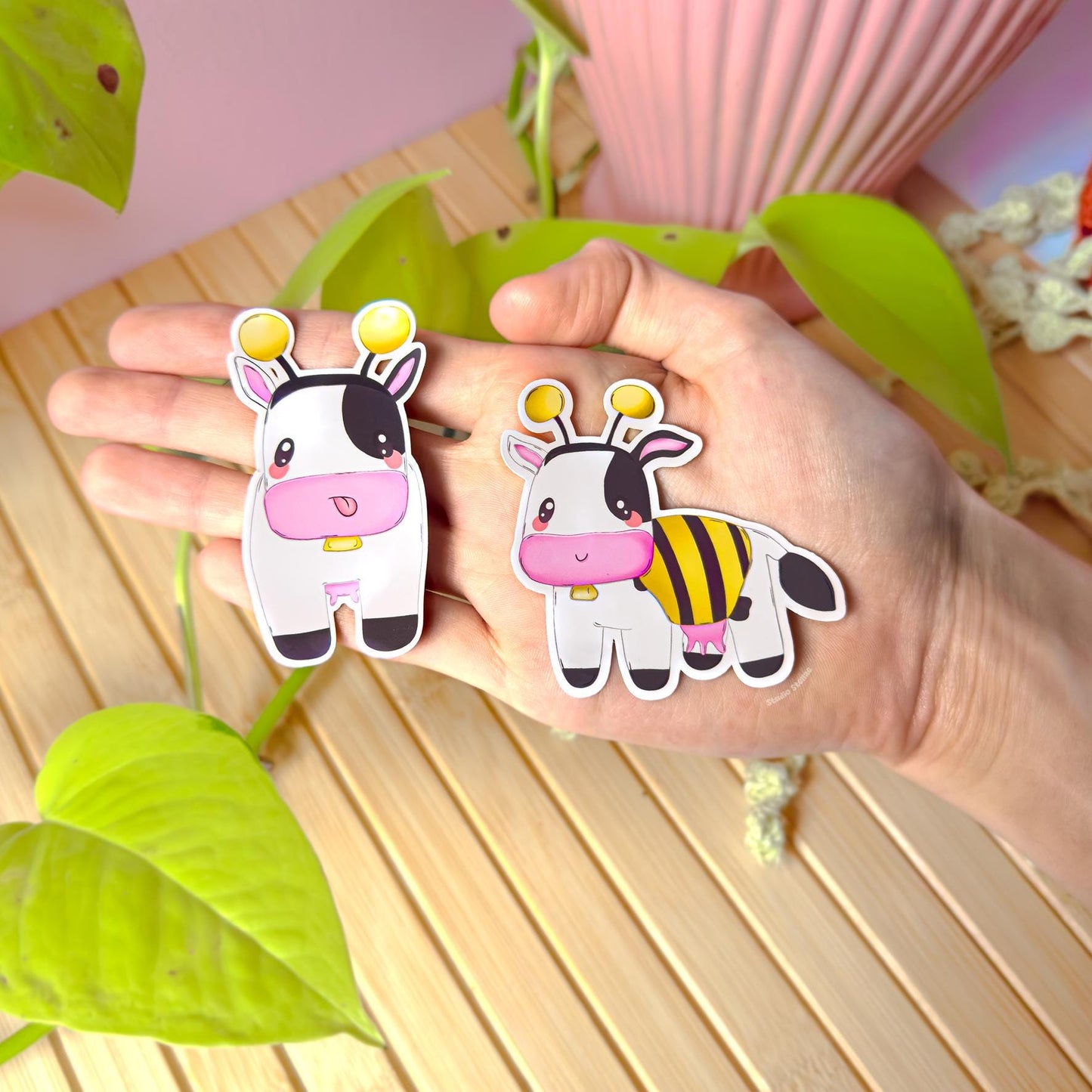 Hand holding both bee cow stickers near green plants and a cozy desk setup, showing real size, colors, and finish in natural light. This lifestyle photo highlights the cute cow sticker set together as a matching pair—perfect for planners, journals, laptops, or water bottles. Great for buyers who want a cozy gamer vibe, farm game nostalgia, or a sweet kawaii sticker pack as a gift.