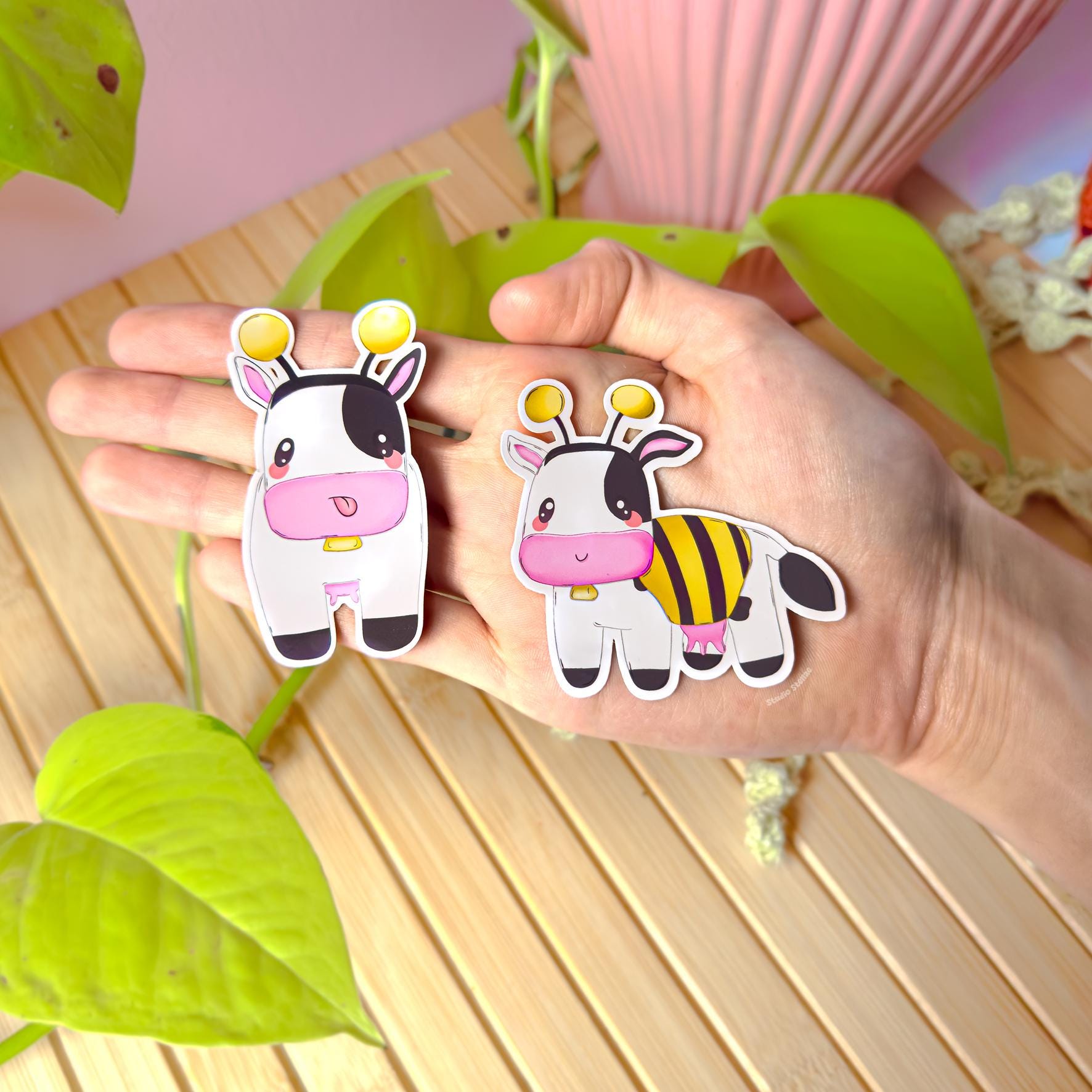 Hand holding both bee cow stickers near green plants and a cozy desk setup, showing real size, colors, and finish in natural light. This lifestyle photo highlights the cute cow sticker set together as a matching pair—perfect for planners, journals, laptops, or water bottles. Great for buyers who want a cozy gamer vibe, farm game nostalgia, or a sweet kawaii sticker pack as a gift.