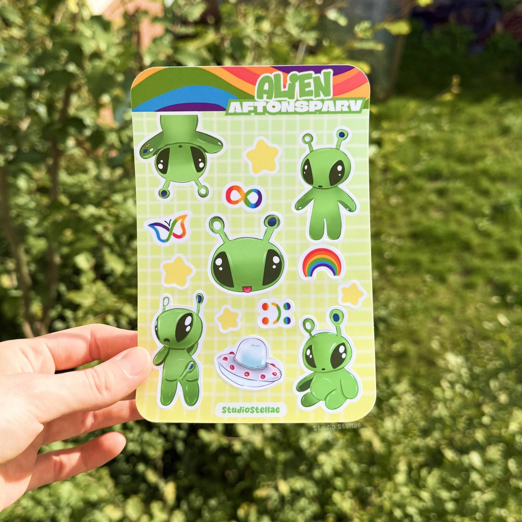 Hand holding the Aftonsparv alien sticker sheet outdoors against a leafy green background, showing the full sticker sheet in natural light for accurate color and size. The kawaii IKEA alien fan art designs include cute green aliens, rainbow details, stars, and neurodivergent symbols that celebrate mental health, ADHD, and autism pride. Perfect for planners, journals, laptops, water bottles, phone cases, and iPads as a supportive neurodivergent gift.