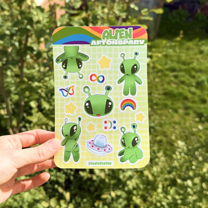 Hand holding the Aftonsparv alien sticker sheet outdoors against a leafy green background, showing the full sticker sheet in natural light for accurate color and size. The kawaii IKEA alien fan art designs include cute green aliens, rainbow details, stars, and neurodivergent symbols that celebrate mental health, ADHD, and autism pride. Perfect for planners, journals, laptops, water bottles, phone cases, and iPads as a supportive neurodivergent gift.