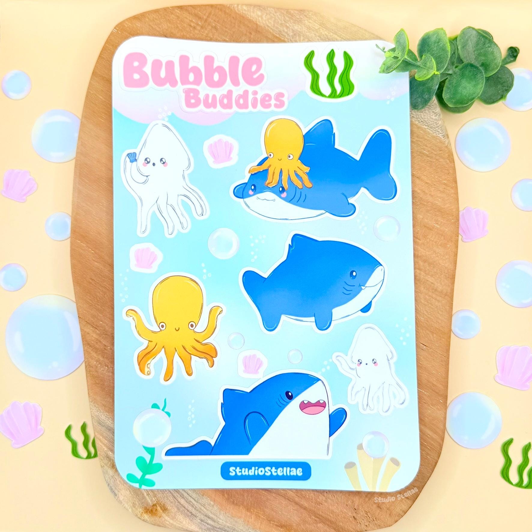 Full Bubble Buddies shark sticker sheet displayed on a soft pastel surface with decorative props, showing the entire collection of cute ocean characters including smiling sharks, a squid, an octopus, bubbles, and tiny seashell details. The photo gives a clear overview of the design, colors, and layout, helping shoppers see exactly what’s included in this kawaii ocean sticker sheet for decorating planners, journals, laptops, or water bottles.