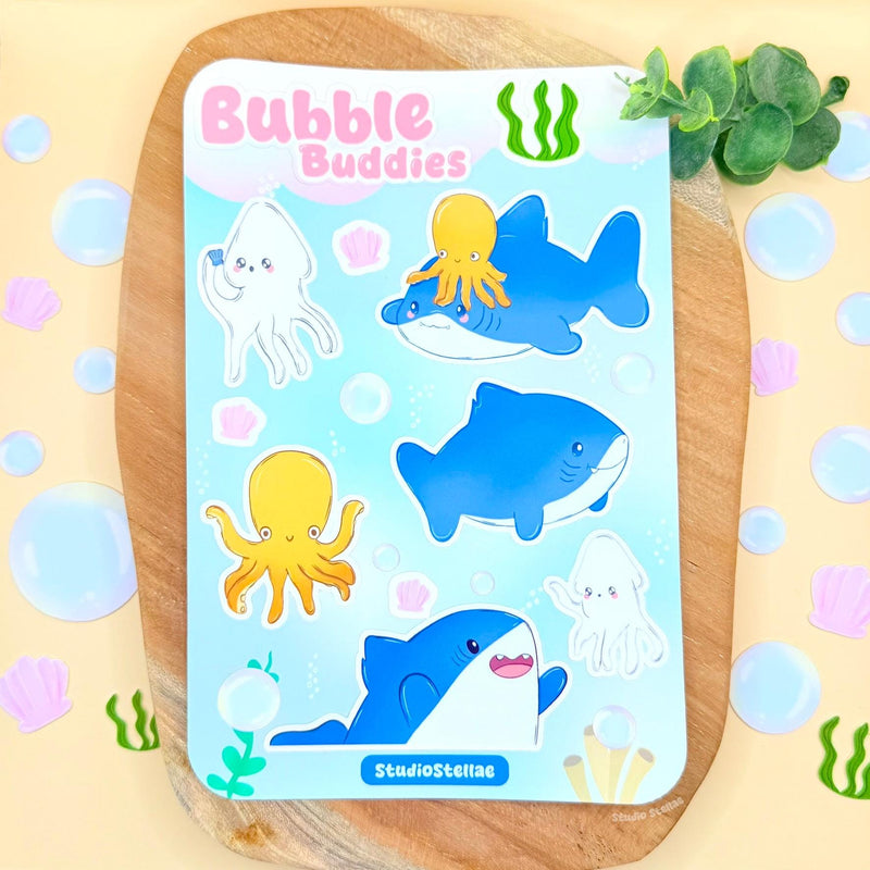 Full Bubble Buddies shark sticker sheet displayed on a soft pastel surface with decorative props, showing the entire collection of cute ocean characters including smiling sharks, a squid, an octopus, bubbles, and tiny seashell details. The photo gives a clear overview of the design, colors, and layout, helping shoppers see exactly what’s included in this kawaii ocean sticker sheet for decorating planners, journals, laptops, or water bottles.