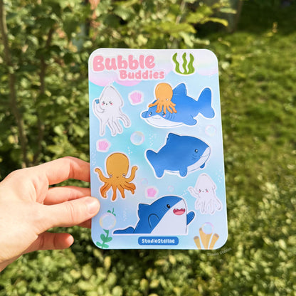 Hand holding the shark sticker sheet outdoors against a leafy green background in natural light, giving an accurate sense of color and size. The ocean characters stand out clearly, helping buyers visualize how the sticker sheet looks in different lighting and reinforcing the playful sea theme.