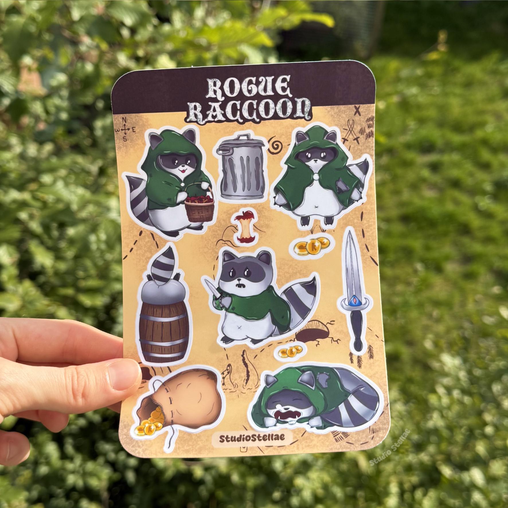 Hand holding the Rogue Raccoon sticker sheet outdoors against a lush green grass background in natural daylight. The full sheet is clearly visible, showing chibi raccoon rogue characters wearing green cloaks alongside fantasy accessories like gold coins, a treasure sack, dagger, barrel, and trash can. This lifestyle photo demonstrates real-life scale, color vibrancy, and how the stickers look in natural light, perfect for DnD players, tabletop RPG fans, journaling, and decorating personal items.