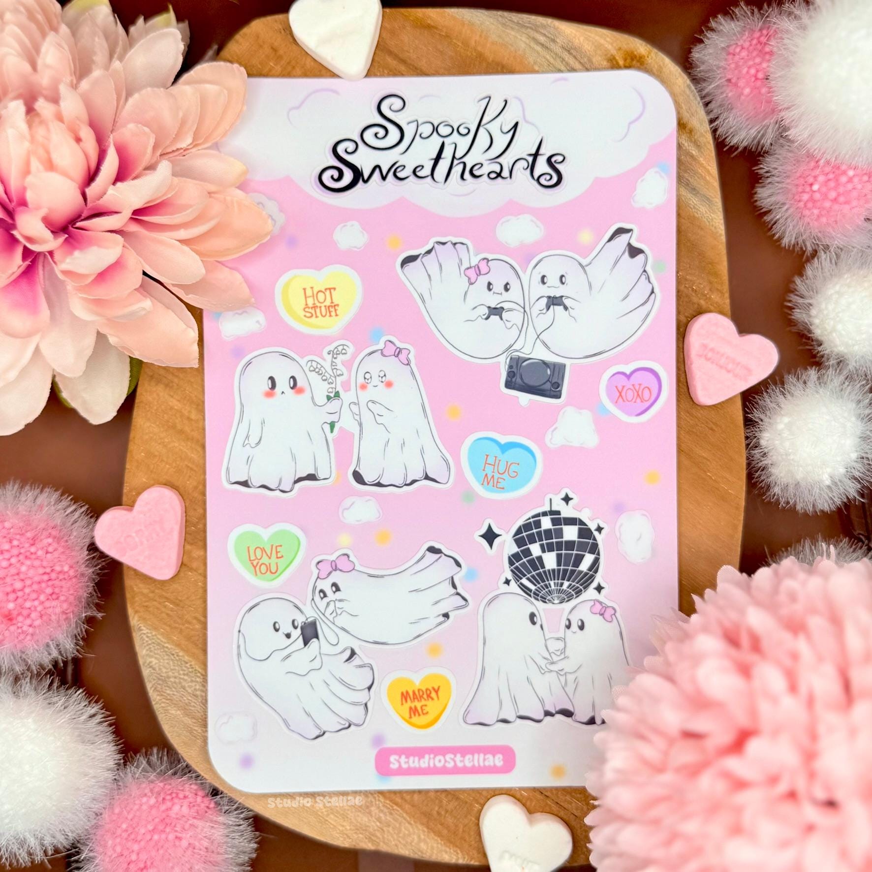 Spooky Sweethearts ghost love sticker sheet styled on a wooden board surrounded by soft pink flowers, fluffy pom poms, and pastel decorations. The sheet features cute ghost couples in romantic scenes like dancing, gaming, hugging, and sharing sweet moments, along with candy heart stickers and pink clouds. The soft pastel pink background and matte laminated finish create a cozy Valentine and cute spooky aesthetic perfect for ghost lovers and romantic sticker collectors.