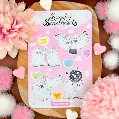 Spooky Sweethearts ghost love sticker sheet styled on a wooden board surrounded by soft pink flowers, fluffy pom poms, and pastel decorations. The sheet features cute ghost couples in romantic scenes like dancing, gaming, hugging, and sharing sweet moments, along with candy heart stickers and pink clouds. The soft pastel pink background and matte laminated finish create a cozy Valentine and cute spooky aesthetic perfect for ghost lovers and romantic sticker collectors.