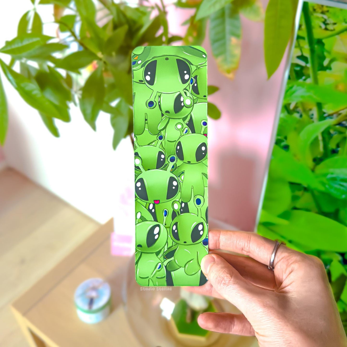 Close-up of the green Aftonsparv-inspired alien bookmark held upright in front of lush green plant leaves. The plushie-style alien is clearly visible, featuring bright green characters with large black eyes and tiny antenna details against a matching green background. The slim rectangular shape, rounded corners, and smooth matte laminated finish are easy to see. This lifestyle image highlights color vibrancy, thickness, and how the bookmark looks in real-world reading settings for book lovers.