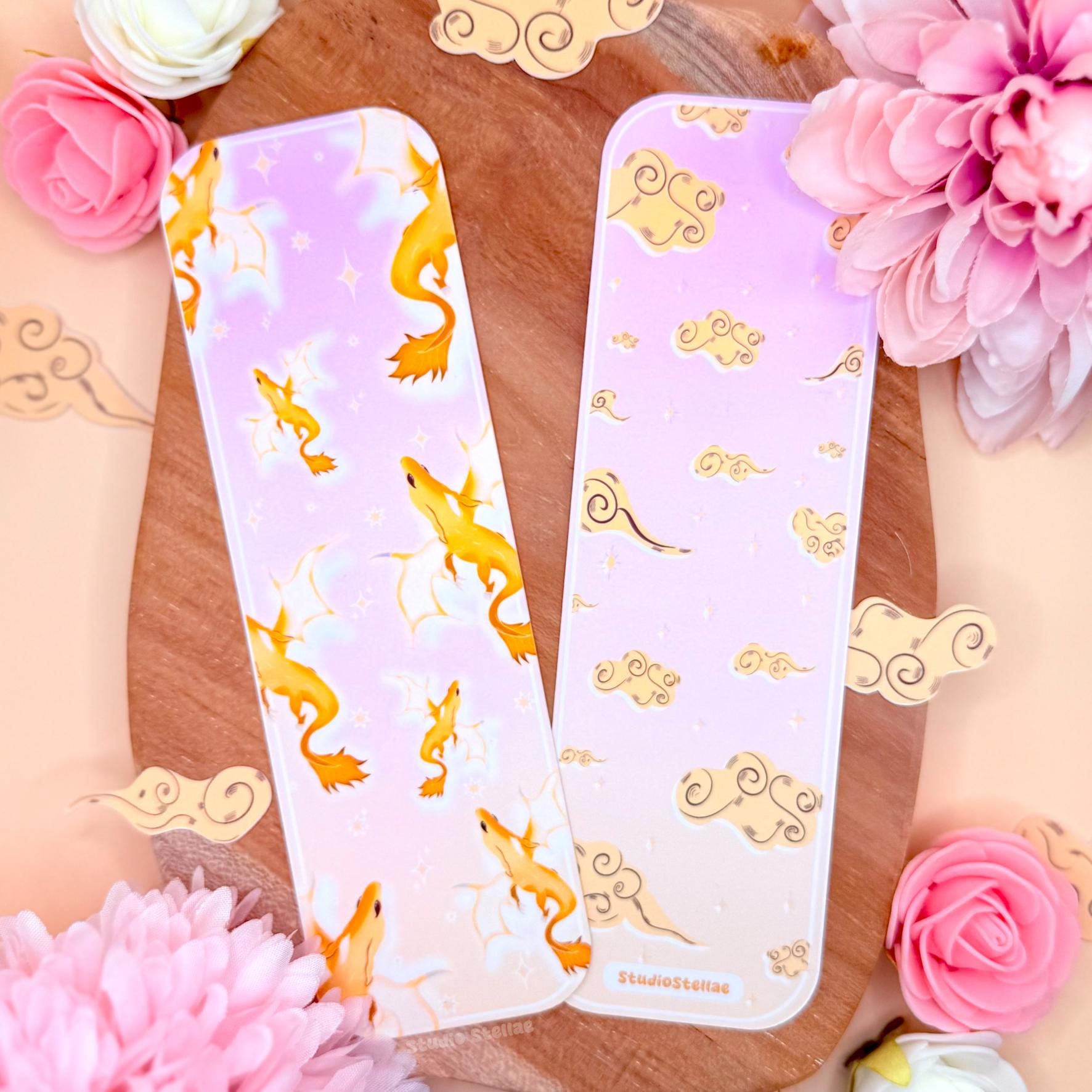 Two pastel Andarna-inspired dragon bookmarks displayed on a wooden surface surrounded by soft pink flowers and delicate decorative accents. The bookmarks feature a golden fantasy dragon illustrated in soft pastel tones with clouds and subtle star details. The matte laminated finish and slim rectangular shape are visible, creating a dreamy romantasy aesthetic perfect for Fourth Wing inspired readers, fantasy lovers, and BookTok collectors.