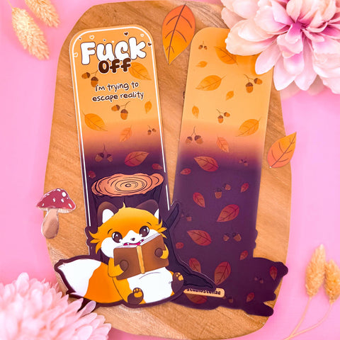 Funny fox bookmark displayed upright against a soft pink background with decorative flowers and cozy autumn accents. The uniquely shaped cottagecore bookmark features a cute orange fox reading a book beside a tree stump, surrounded by forest leaves and acorns. The top text reads “Fuck Off I’m trying to escape reality,” adding humorous introvert energy. The matte laminated finish and detailed forest illustration create a cozy book lover aesthetic perfect for readers and cottagecore fans.